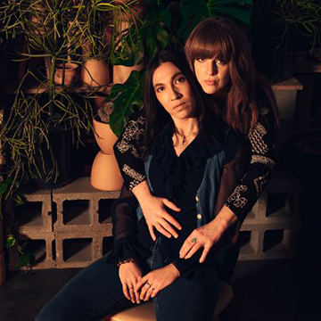 two women - one is sitting in a chair and the other stands behind with arms around her