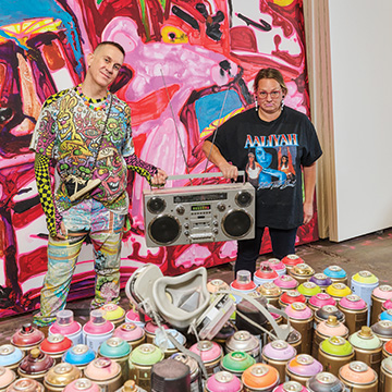 Jeremy Scott and Katherine Bernhardt standing next to each other in an art studio. They are holding a boom box between them and standing in front of a large, colorful painting and spray paint bottles.