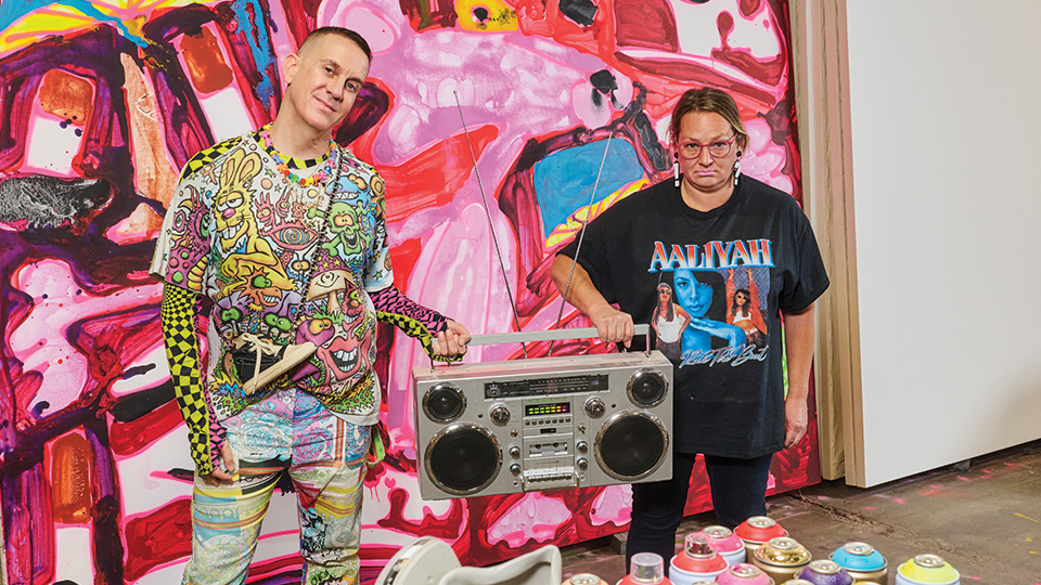 Jeremy Scott and Katherine Bernhardt standing next to each other in an art studio. They are holding a boom box between them and are standing in front of a large, colorful painting.