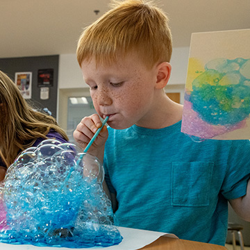 three young children blow dyed bubbles onto paper to make an abstract work of art