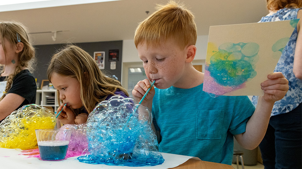 three young children blow dyed bubbles onto paper to make an abstract work of art