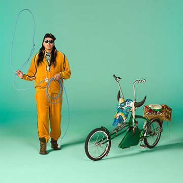 print by Dana Claxton depicting a man with long black hair, sunglasses and hat wearing a yellow track suit. He is standing emotionless next to a bicycle decorated with a cow skull while holding up a blue lasso.