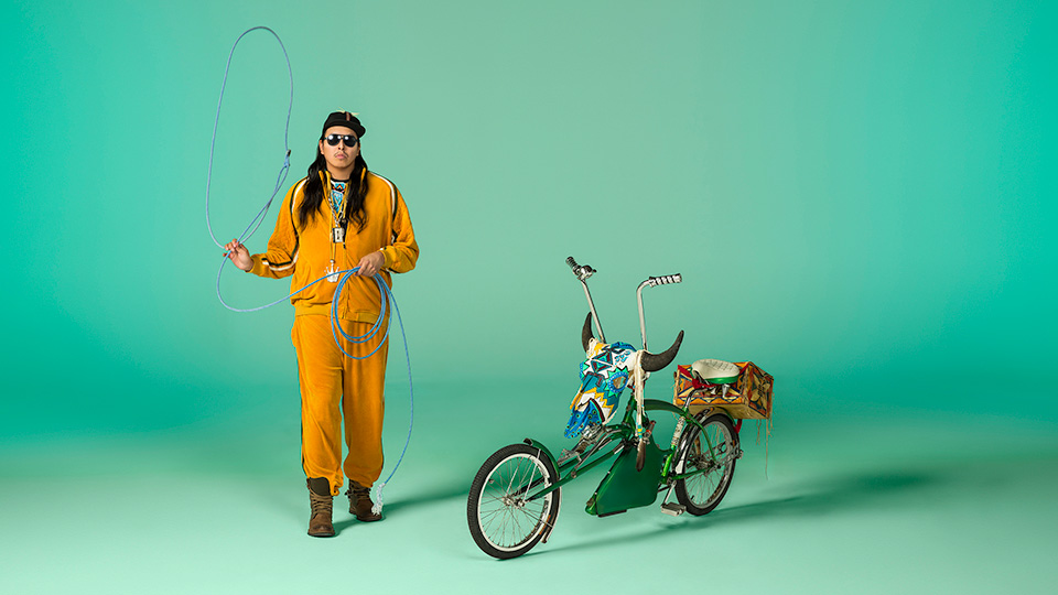 print by Dana Claxton depicting a man with long black hair, sunglasses and hat wearing a yellow track suit. He is standing emotionless next to a bicycle decorated with a cow skull while holding up a blue lasso.