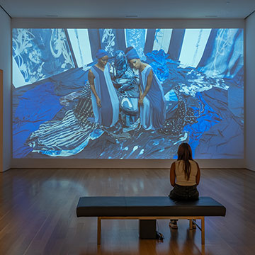 A woman sits on a bench and watches a large projection taking up the entire wall in front of her. The projection is an overhead view of two women wearing blue garments surrounded by blue fabric.