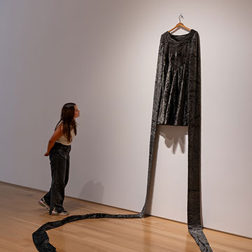 A woman peers at an art installation at the Nerman. The work is a black garment hanging on the wall. The sleeves of the garment form a continuous line; they reach to the floor and form a circle on the floor.