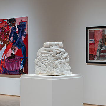 Three works on display at the Nerman. To the left is a brightly colored abstract painting of a figure. In the center is a white sculpture that appears to be tubes or forms folded and stacked on each other. To the right is another brightly colored painting depicting two figures sitting on a building stoop.