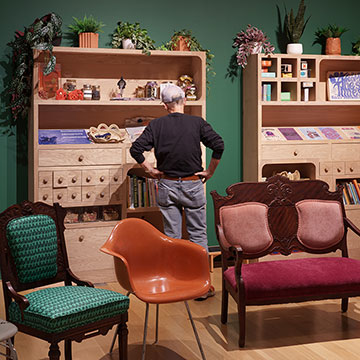A man stands between assorted chairs and a bookshelf. He has his back to the camera, his hands on his hips, and is looking at the bookshelf.
