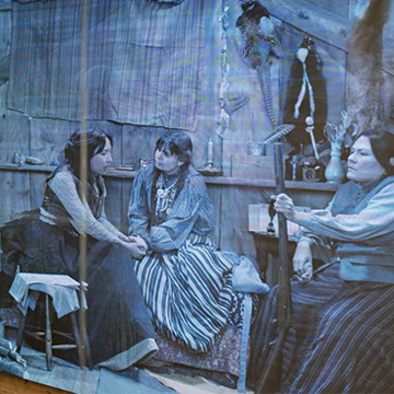 3 Native American women sitting in a wooden structure. There is a wood burning stove next to them and animal skins on the wall. One woman is holding a shotgun.