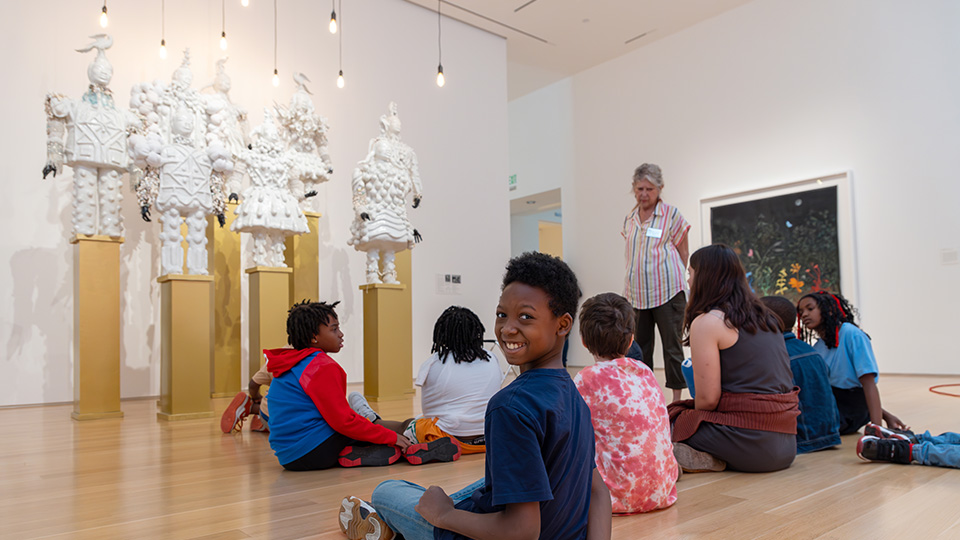 a boy sitting criss-cross on the floor in front of a sculpture turns back to smile at the camera. He sitting with a group of children on a tour at the Nerman.
